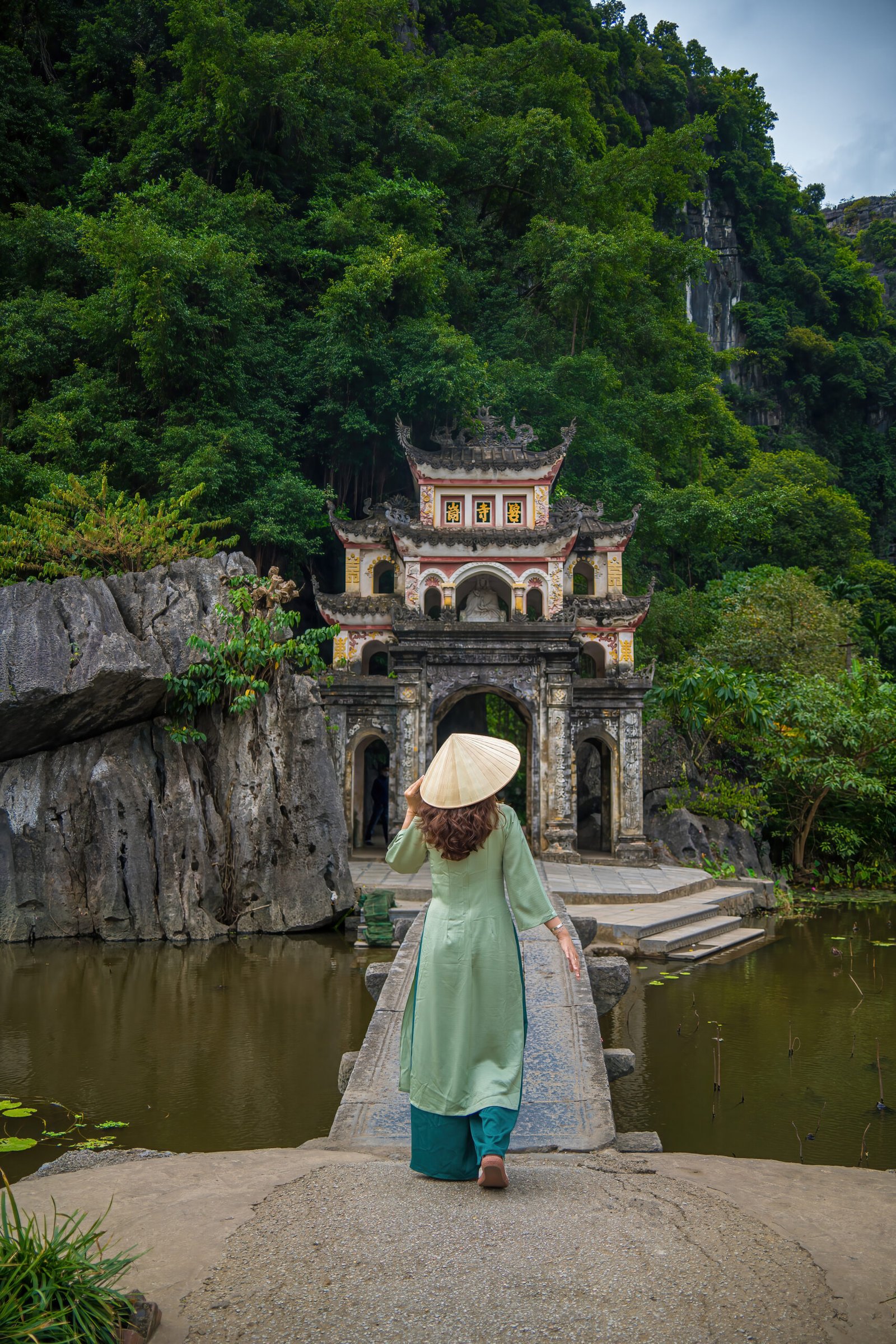 back view of girl wearing ao dai in front of outdoor park landscape with lake and stone bridge. gate entrance to ancient bich dong pagoda complex. ninh binh, vietnam travel destination